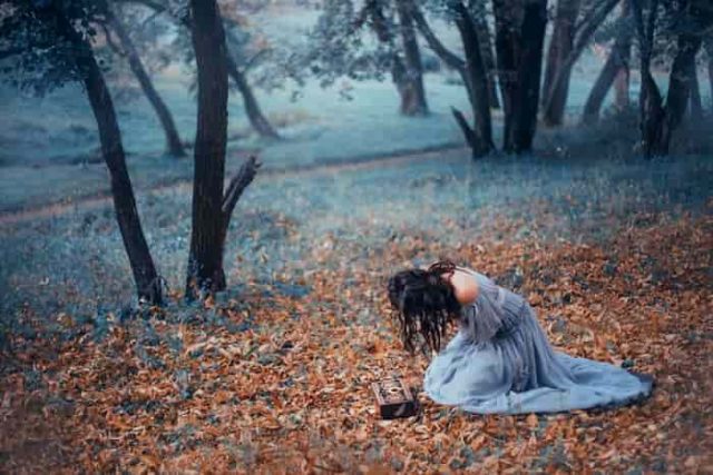 Ελπίδα. Μελαχρινή κοπέλα σε ένα μακρύ, ανοιχτό γκρι, ντεμοντέ φόρεμα, κάθεται στο κρύο έδαφος που καλύπτεται με Φθινοπωρινά πεσμένα φύλλα, σε ένα σκοτεινό δάσος, κλαίει πάνω από το ανοικτό κουτί της Πανδώρας