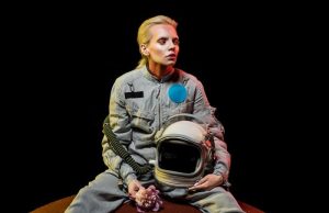 ΑΡΚΤΟΥΡΙΑΝΟΙ, ΤΕΛΟΣΙΑΝΟΙ, ΛΥΡΙΟΙ, ΑΓΓΕΛΟΙ γυναίκα με διαστημική στολή, κρατάει κράνος και λουλούδι, κάθεται σε ένα πλανήτη